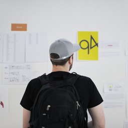 A man wearing a baseball cap standing in front of a whiteboard filled with paper sheets